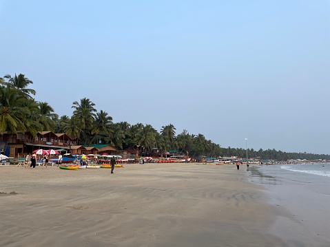 Palolem Beach, Goa, India - November, 29 2022: Stock photo showing row of beach hut shacks under tropical coconut palm trees on Palolem Beach, Goa, India. Sunbathers on holiday vacation sun loungers, holidaymakers swimming in sea, kayaks, canoes, fishing boats and parasol umbrellas.