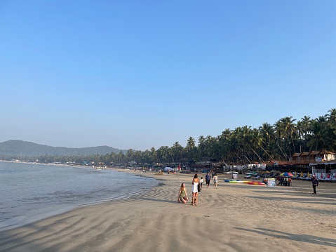Palolem Beach, Goa, India - December, 1 2023: Stock photo showing row of beach hut shacks under tropical coconut palm trees on Palolem Beach, Goa, India. Sunbathers on holiday vacation sun loungers, holidaymakers swimming in sea, kayaks, canoes, fishing boats and parasol umbrellas.