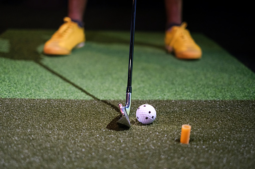 Close up shot of unrecognizable man playing golf indoor in simulator. The man feet standing a step behind the golf club and tee.