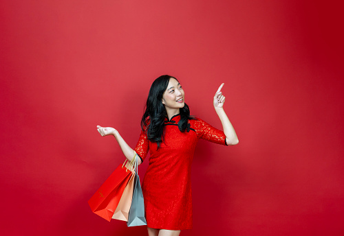 Asian chinese woman with red cheongsam or qipao holding shopping bag wish the good luck sale and prosperity in Chinese New Year celebration holiday isolated on the red background