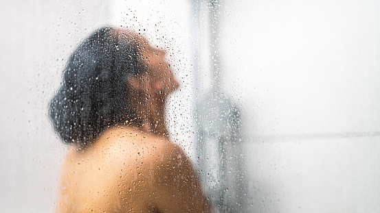 Woman with black hair showering, view behind fogged glass