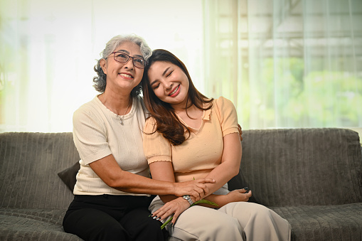 Smiling grey haired elderly woman embracing her grownup daughter sitting on sofa at home.