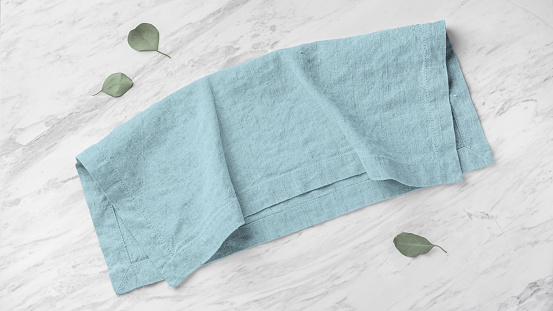 Blue linen napkin folded on marble kitchen table with green leaves, top view