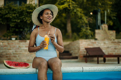 Mature African American woman, relaxed by the pool, enjoying a fruity drink and a delicious fruits