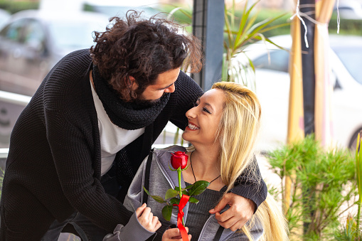 Young man hugging his girlfriend from behind and surprising her with a  red rose.