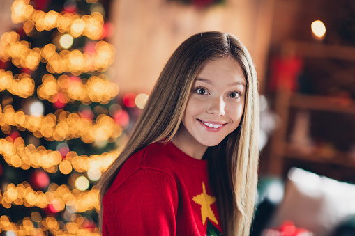 Portrait of girl teenager in red jumper toothy smile during lights on christmas tree lightning as decoration with home interior background.