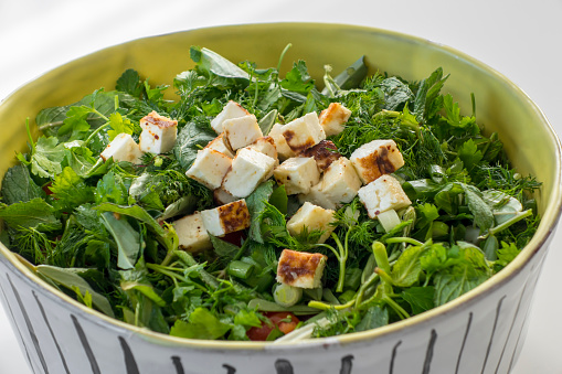 Mixed herb salad with fried halloumi cheese. It contains Halloumi cheese, 1 Bunch of Mixed Endemic Island Herbs, 2 Lettuce Leaves, 1 Spring Onion and 1 Fresh Garlic. Close-up in a large handmade ceramic bowl and on a white background. No people.