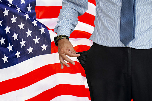 Close-up of hand of man in suit turning pocket of his trousers inside out and showing it empty on the background of the USA flag. Crisis, unemployment, bankruptcy concept in US