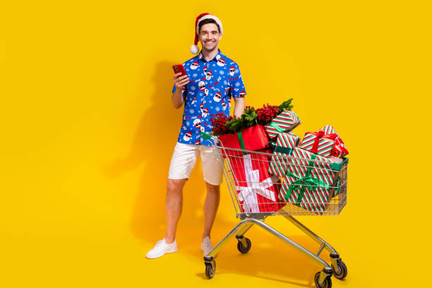 istock Full size photo of cheerful handsome guy use smart phone pile stack x-mas giftbox trolley isolated on yellow color background 1812967757