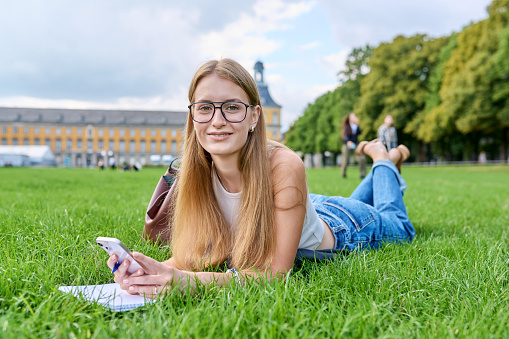 Portrait of young teenage girl student lying on lawn on grass, using smartphone writing in notebook, looking at camera, educational building background. Youth, education, college university concept