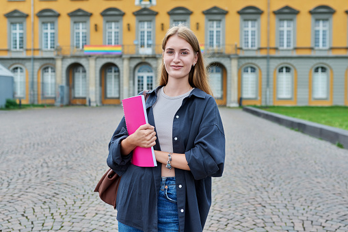 Portrait of young teenage girl student posing outdoors, background of educational building. Smiling female with bag, textbook looking at camera. Youth, education, college, university