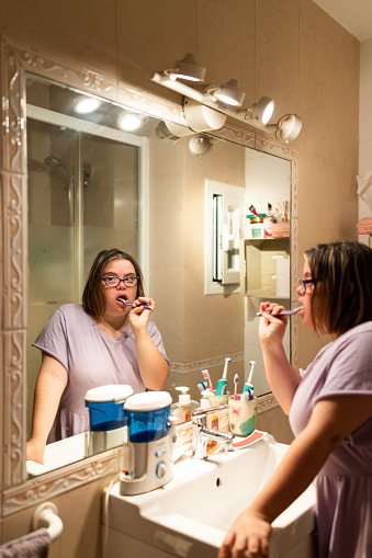 Vertical photo of the image reflected on a mirror of a woman with down syndrome cleaning her teeth in the bathroom