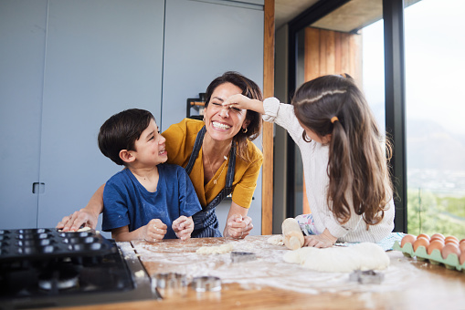 Mother and her two cute young children laughing and having fun with flour while making cookie dough together at a counter in their kitchen at home