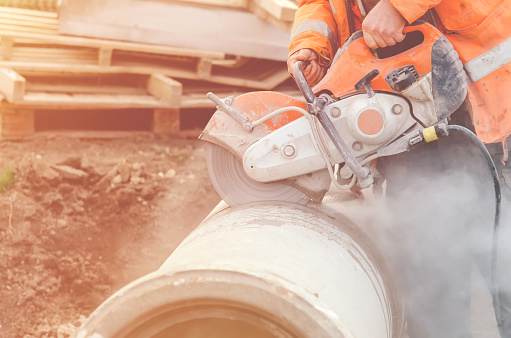 A worker at the construction site cutting a concrete drainage pipe with a petrol concrete saw. Builder covered in a hazardous dust cloud as safety procedure breached on construction site.