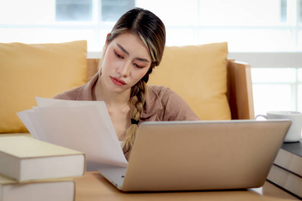 istock Serious tired beautiful woman reads many document papers, analyzes data information, uses laptop computer. Thoughtful confused young female student feels stressed while studying of a test at home. 1813132022