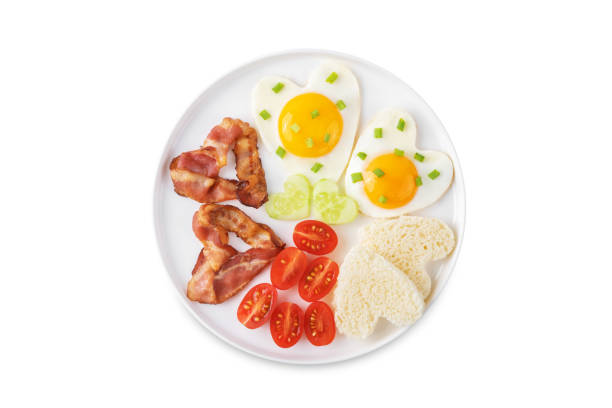 Fresh breakfast with fried eggs, fried bacon slices, bread slices and vegetables in the form of heart for Valentine's day holiday on a white isolated background