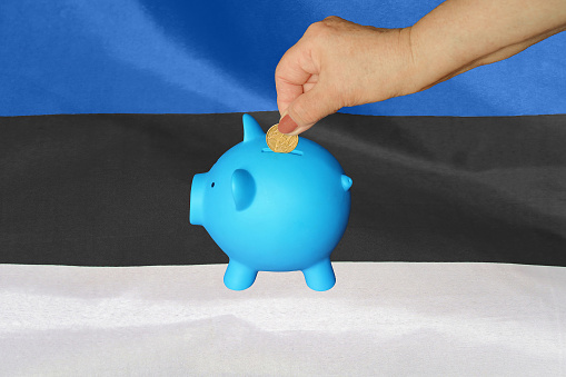 Hand of elderly woman putting coin into piggy bank on Estonia flag background. Hand putting coin to piggy bank. Retirement savings, piggy bank. Concept saving money and retirement fund in Estonia