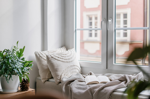 Cozy corner on windowsill in nordic style room in flat with big windows and view on city buildings. Place for reading books at home. Beige textile and pillows near white wall.