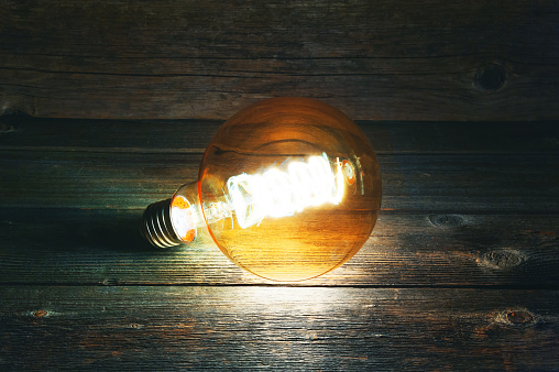 A decorative burning light bulb lies without a connection on old wooden boards in the dark. Side view.