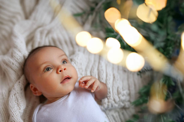 新年のクリスマス休暇のシーン - 面白いかわいい新生児興奮した男の子は、クリスマスのお祝いツリーの金のおもちゃとライトの花輪を見てニットの毛布の上に横たわっています。幼児の子� - christmas textile blanket decoration ストックフォトと画像