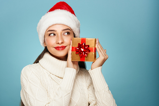 Beautiful in a white knitted sweater and santa hat with gift box isolated on blue background. Christmas or New Year festivities. Holiday and Party