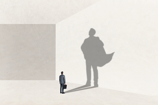 A man stands while holding his briefcase as he looks up at his shadow that is cast on the wall in front of him that shows imr wearing a cape and expressing power and confidence.