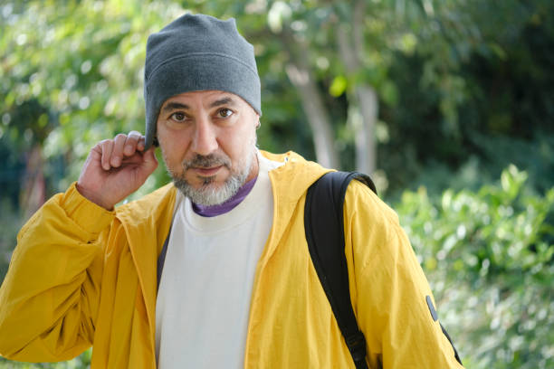 A mature man is looking at the camera on the public park. He carries a bag on his shoulder.