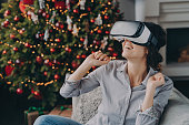 Amazed woman wearing virtual reality glasses first time, sitting on sofa at home near Christmas tree