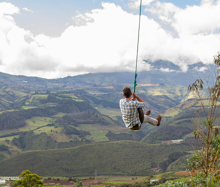 A local backpacker tourist male enjoying the landscape view swinging on top of the Pichincha's volcano mountain. On Back, Quito's ecuadorian capital.

He's wearing a red jacket, grey jeans and blue sunglasses.
