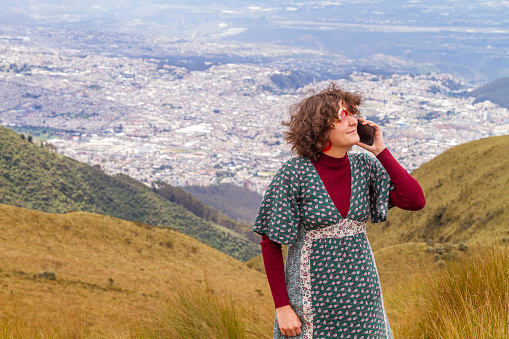 Portrait of young teenager solo traveler female talking with her smartphone with 5G Technology at the top of the Pichincha's volcano mountain. On Back, the ecuatorian capital: Quito.

Carefree backpacker fashion woman, in Quito, Pichincha, Ecuador