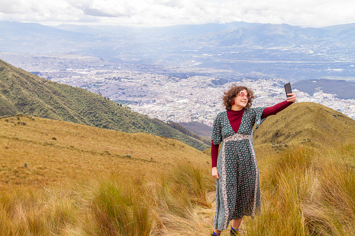 Portrait of young teenager solo traveler female taking a selfie with her smartphone with 5G Technology at the top of the Pichincha's volcano mountain. On Back, the ecuatorian capital: Quito.

Carefree backpacker fashion woman, in Quito, Pichincha, Ecuador.