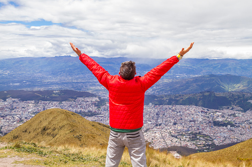 Portrait of young male solo traveler backpacker \nenjoying the trekking and opening his arms on top of the Pichincha's volcano mountain. On Back, Quito's ecuadorian capital.\n\nHe's wearing a red jacket, grey jeans and blue sunglasses.