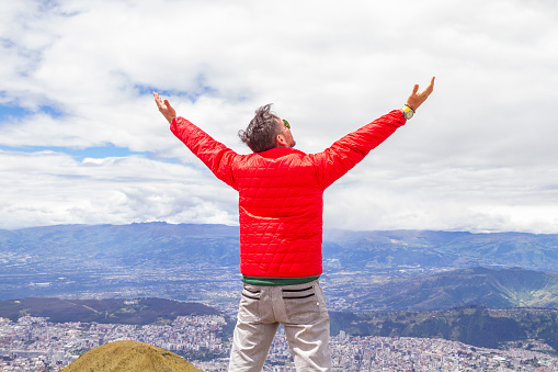 Portrait of young male solo traveler backpacker 
enjoying the trekking and opening his arms on top of the Pichincha's volcano mountain. On Back, Quito's ecuadorian capital.

He's wearing a red jacket, grey jeans and blue sunglasses.