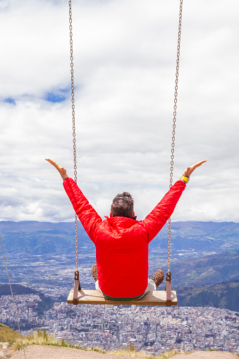 A local backpacker tourist male enjoying the landscape view swinging on top of the Pichincha's volcano mountain. On Back, Quito's ecuadorian capital.\n\nHe's wearing a red jacket, grey jeans and blue sunglasses.