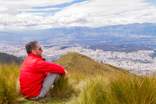 Portrait of young male solo traveler backpacker 
enjoying the trekking and opening his arms on top of the Pichincha's volcano mountain. On Back, Quito's ecuadorian capital.

He's wearing a red jacket, grey jeans and blue sunglasses.