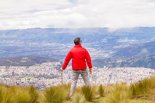 Portrait of young male solo traveler backpacker \nenjoying the view finishing his trekking activity on top of the Pichincha's volcano mountain. He is standing in front of Quito's ecuadorian capital.\n\nHe's wearing a red jacket, grey jeans and blue sunglasses.