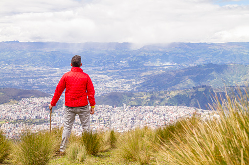 Portrait of young male solo traveler backpacker 
enjoying the view finishing his trekking activity on top of the Pichincha's volcano mountain. He is standing in front of Quito's ecuadorian capital.

He's wearing a red jacket, grey jeans and blue sunglasses.