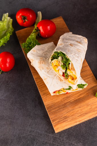 Tortilla wrapped with fried chicken meat, vegetables and corn. Tasty cut burrito on dark background