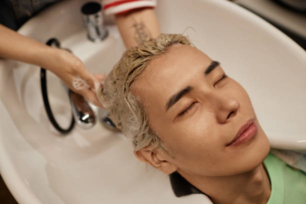 asiatischer mann genießt haarwäsche und haarpflegebehandlung im waschbecken des schönheitssalons - lightener stock-fotos und bilder