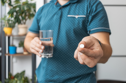 Man taking medicine, holding in a hand white therapeutic pill, antibiotics, painkiller and glass of water, close-up view.