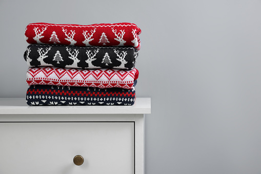 Different folded Christmas sweaters on chest of drawers against grey background. Space for text
