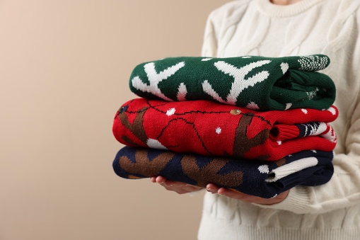 Woman holding stack of different Christmas sweaters on beige background, closeup. Space for text