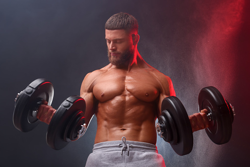 Young bodybuilder exercising with dumbbells in smoke on color background, low angle view