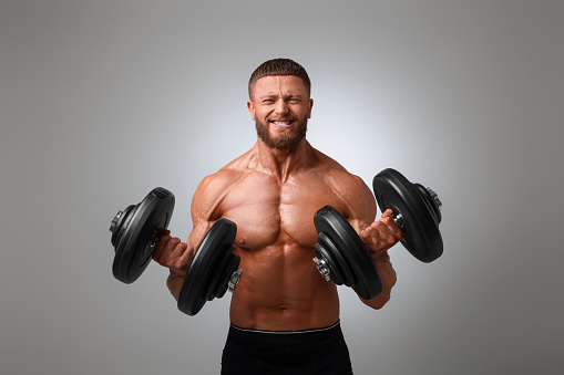 Emotional young bodybuilder exercising with dumbbells on light grey background