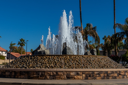 Large water fountain surrounded by rock wall and ledge, palm trees in background