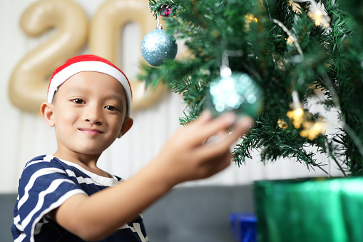 Merry Christmas 2024 concept Asian boy decorates Christmas tree with colorful Christmas balls.