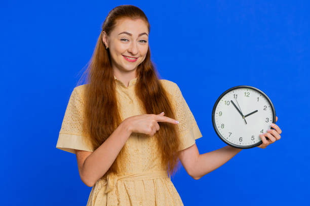 rudowłosa dorosła dziewczyna pokazująca czas na ścianie zegar biurowy, ok, kciuk w górę, zatwierdź, termin się pospiesz - checking the time time clock women zdjęcia i obrazy z banku zdjęć