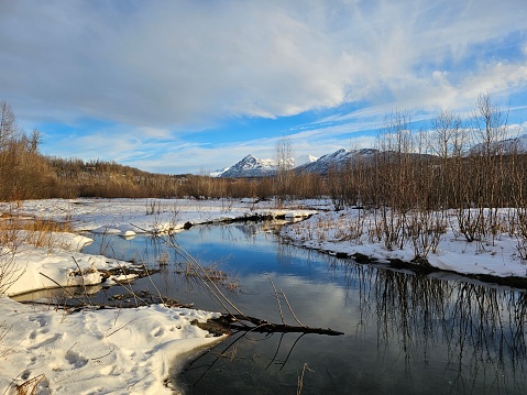 Idyllic winter scene of bright blue snowmelt river, snow covered mountain landscape and bright blue sky with dramatic cloud formation