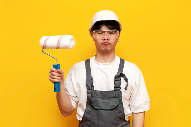 istock puzzled male asian painter in uniform with paint roller is bored on yellow isolated background 1813880694
