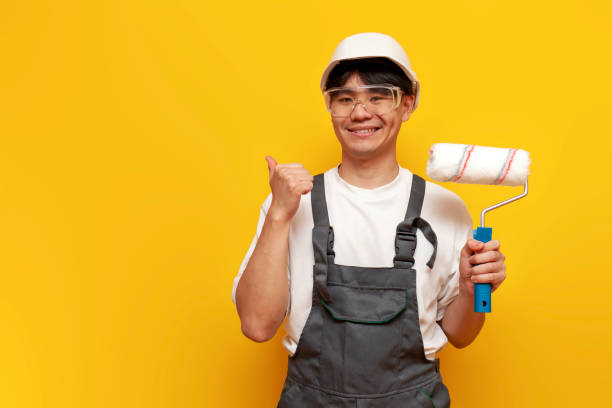 painter asian man in uniform holds roller and points to copy space, korean repairman in hardhat and overalls with tool